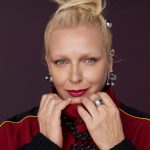 Portret blond kobiety w czerwono-czarnej kurtce, która prezentuje wyrazistą biżuterię jubilerską – wiszące kolczyki, liczne pierścionki na dłoniach oraz kolczyki w uchu.
