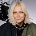 Portret blond kobiety w ciemnym ubraniu z ozdobnym, kulistym naszyjnikiem na szyi, pozującej na jasnym tle z abstrakcyjnymi elementami.
