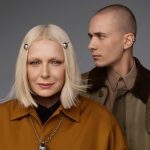 Portret blond kobiety z ozdobnymi spinkami we włosach i srebrnym naszyjnikiem oraz młodego mężczyzny z kolczykiem w uchu na neutralnym tle.