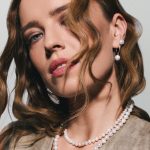 Portret młodej kobiety z jasnobrązowymi, falowanymi włosami, prezentującej elegancką biżuterię z perłami – naszyjnik z pereł oraz pasujące kolczyki z perełkami.