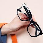 Zdjęcie przedstawiające dłoń trzymającą eleganckie, duże czarne okulary korekcyjne w grubych oprawkach na jasnym tle.