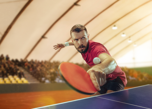 Zdjęcie przedstawiające sportowca w czerwonej koszulce grającego w tenis stołowy podczas zaciętego meczu na hali z publicznością.